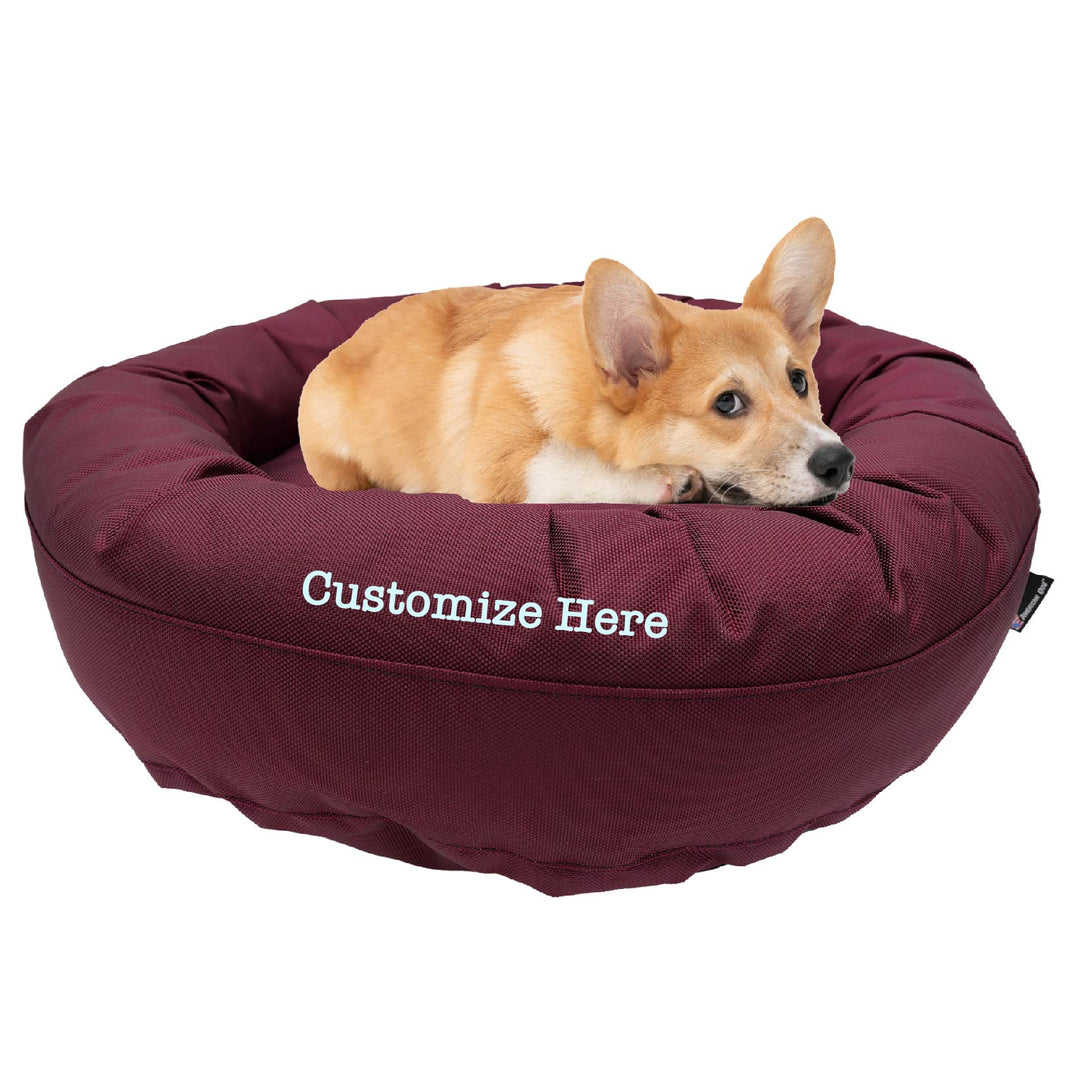 Maroon round bolstered dog bed with a Corky laying in it with 'Customize Here' embroidered on the side.