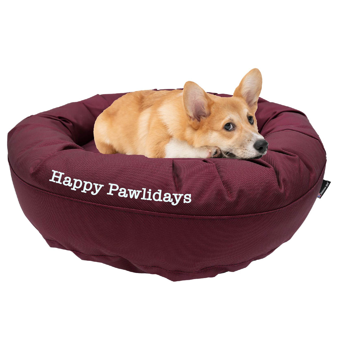 Maroon round bolstered dog bed with a Corky laying in it with 'Happy Pawlidays' embroidered on the side.